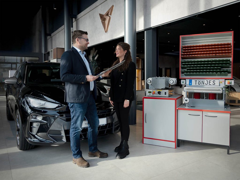 Autohaus-Verkäufer überreicht Kundin frisch gedrucktes Kfz-Kennzeichen.